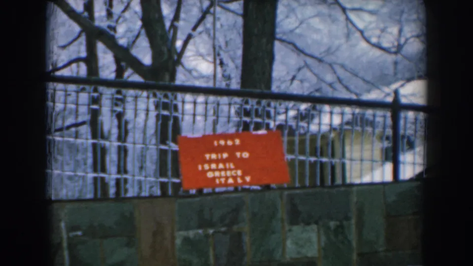 1962 Greece travel-film frame showing a red “TRIP TO ISRAEL GREECE ITALY” sign behind a wire fence and stone wall.