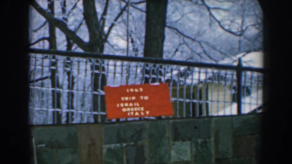 1962 Greece travel-film frame with the red itinerary sign centered behind fencing, trees, and stonework.