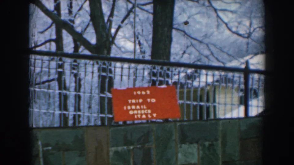1962 Greece travel-film frame showing the red trip sign from a slightly shifted handheld angle behind a fence.