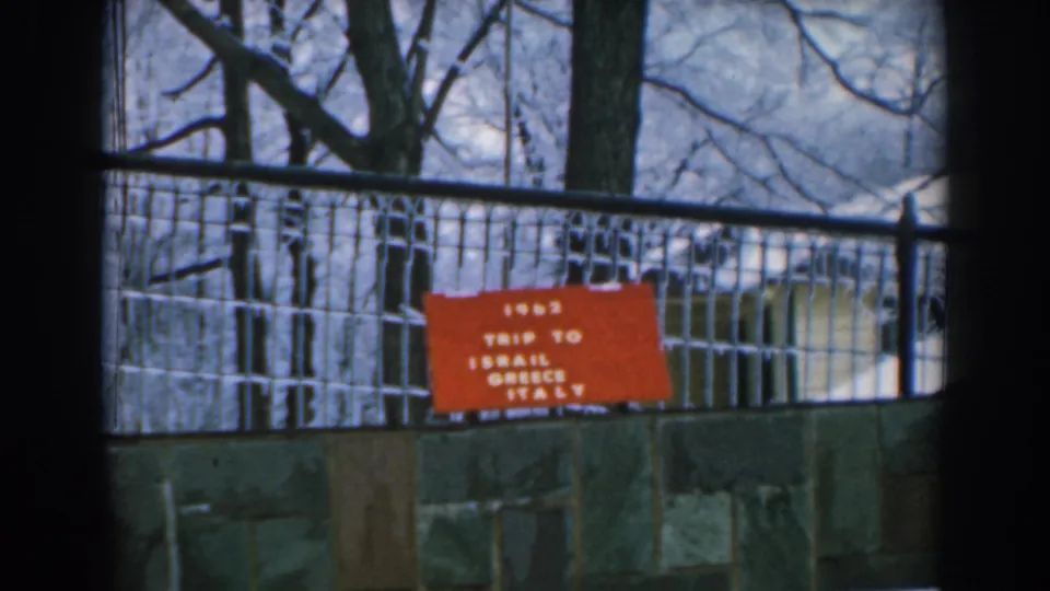 1962 Greece travel-film frame with the red travel sign partly softened by focus and framed by fence rails and trees.