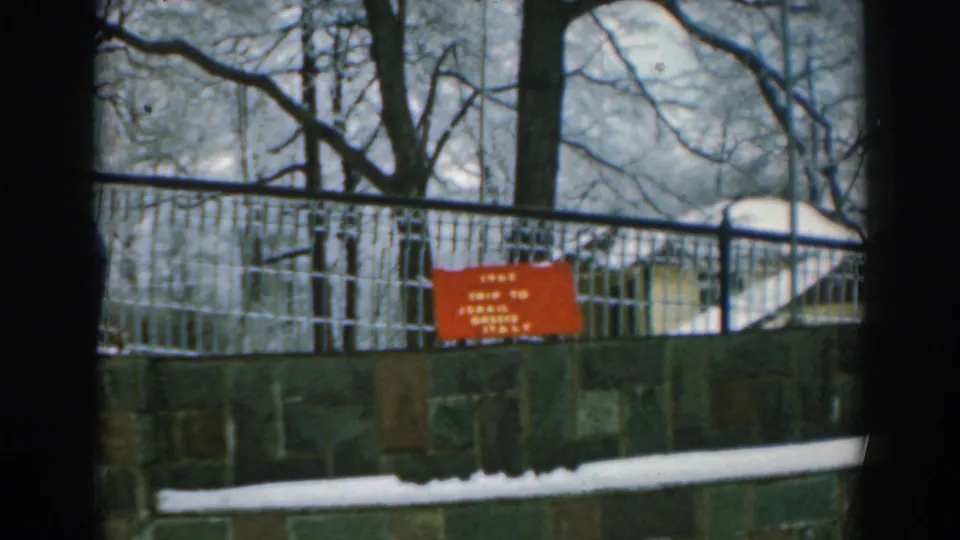 1962 Greece-related home-movie frame of a red travel sign on a snowy exterior wall behind a metal fence and winter trees.