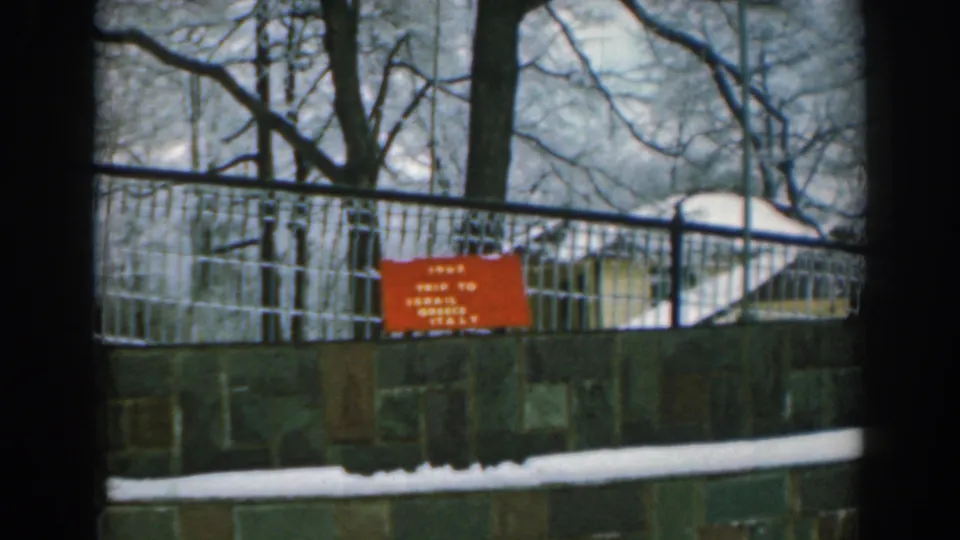 A 1962 8mm travel frame in Greece showing the red trip sign, wire fence, grey wall, snow, and leafless trees.