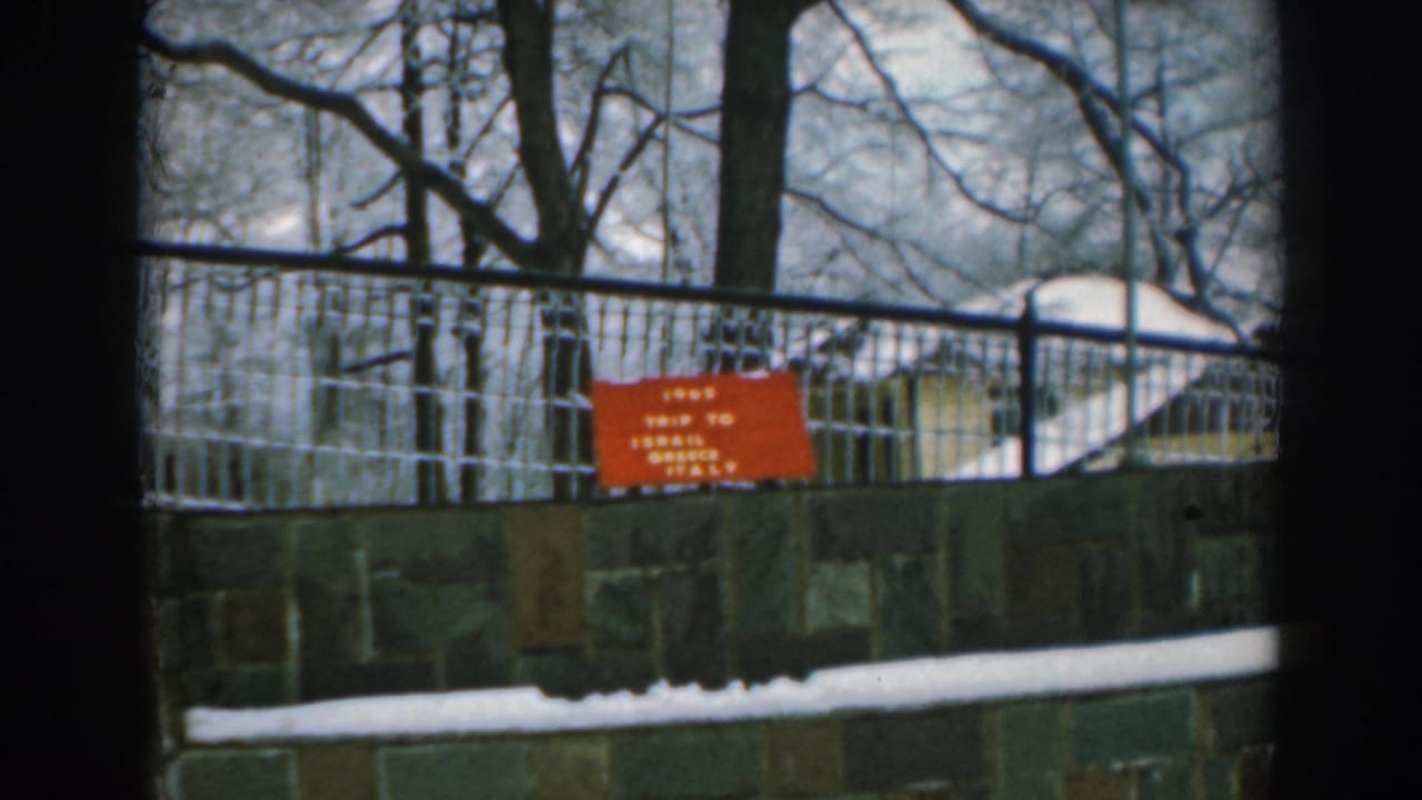 1962 Greece travel-home-movie frame showing a snow-covered park edge, metal railing, stone wall, bare trees, and a red trip sign.