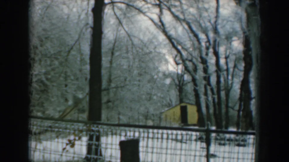 1962 Greek winter view through a window, showing snow-covered trees, wire fencing, and a small yellow shed beyond.