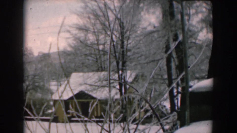 1962 Greece home-movie frame with rustic snow-covered roofs, bare trees, and wooded hills in soft winter light.