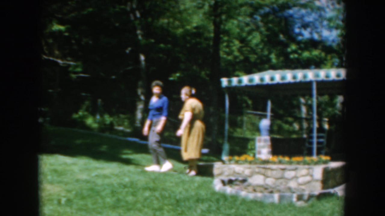 Two women walk across a sunny green lawn beside a stone planter and small roofed garden feature.