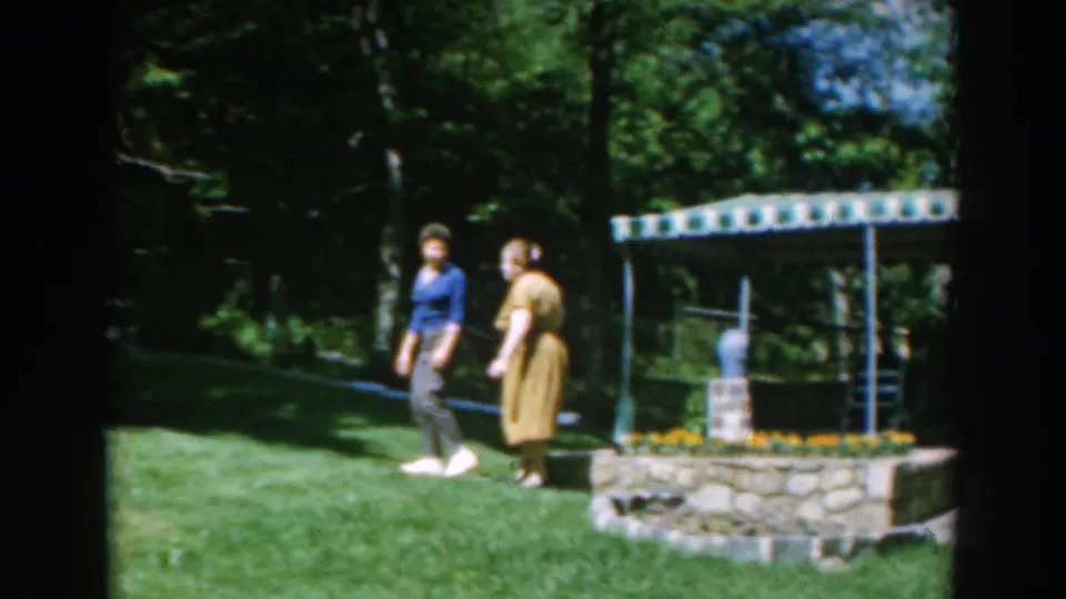 In 1962 Greece home-movie footage, two women walk through a leafy backyard near a stone planter and garden shelter.