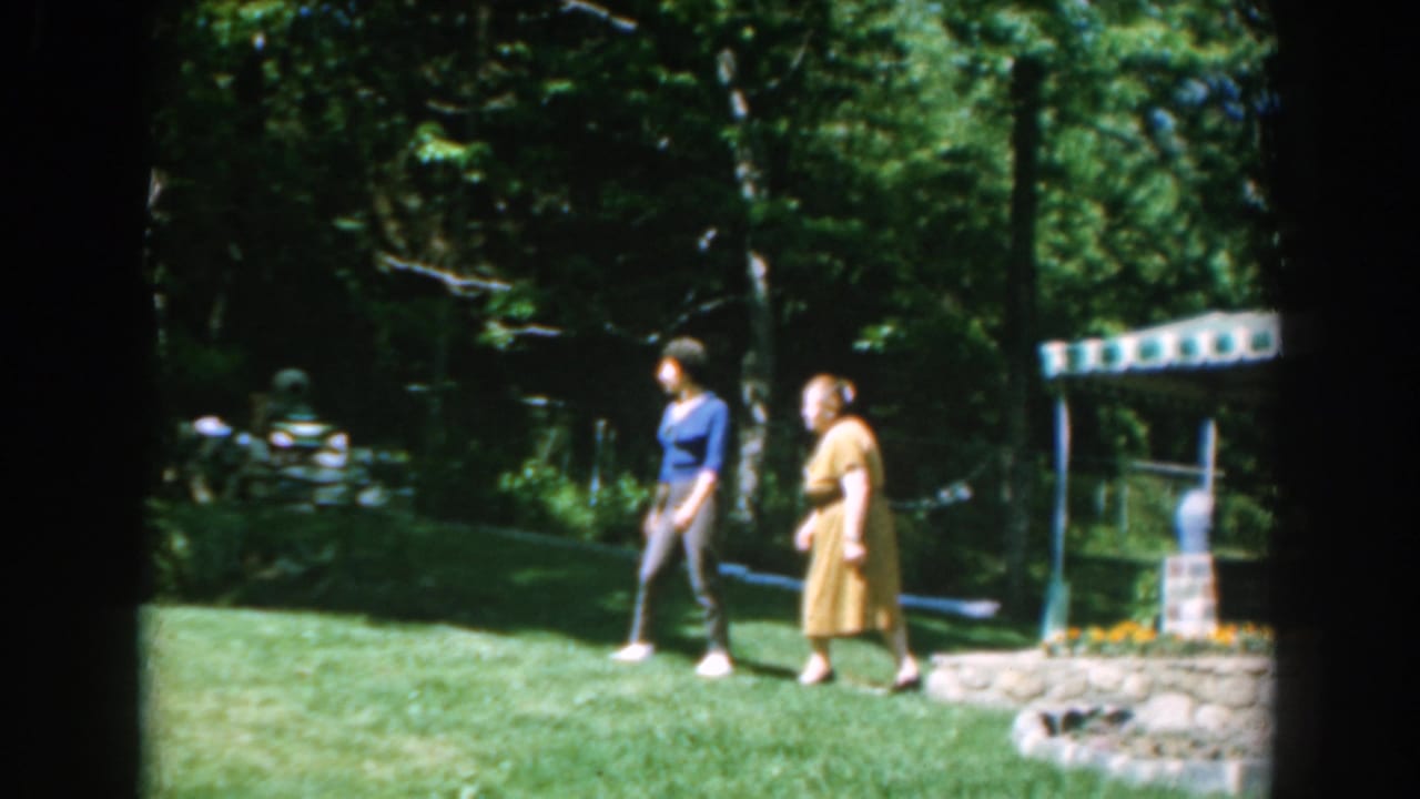 The pair walks through the sunlit garden in 1962 Greece footage, passing a low stone planter and shaded trees.
