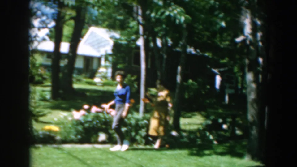 In the garden scene, the women pass a flower bed while a house and trees appear behind them in Greece home-movie footage.