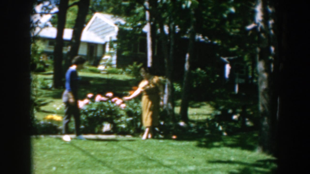 Two women stand near a flower bed in a sunny garden with a white house behind trees.