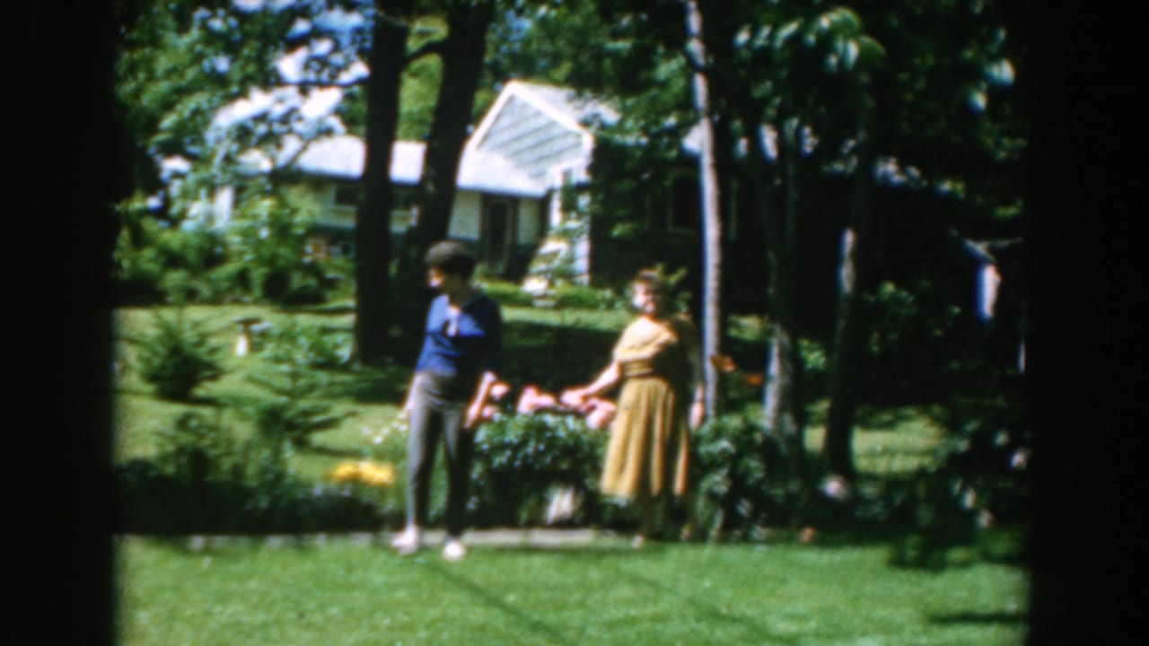 Two women in a 1962 Greece garden scene stand near pink flowers, lawn, trees, and a white house.