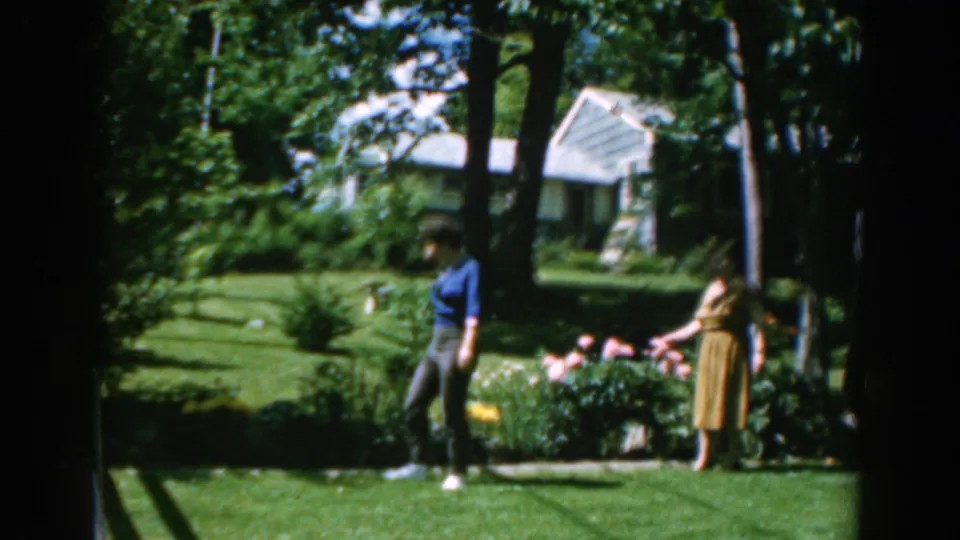 A woman walks across a sunny garden in 1962 Greece while another stands near flowers and shrubs.