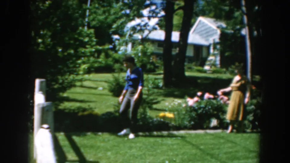Two women remain in a sunlit 1962 Greece garden, one on the lawn and one beside the flower bed.