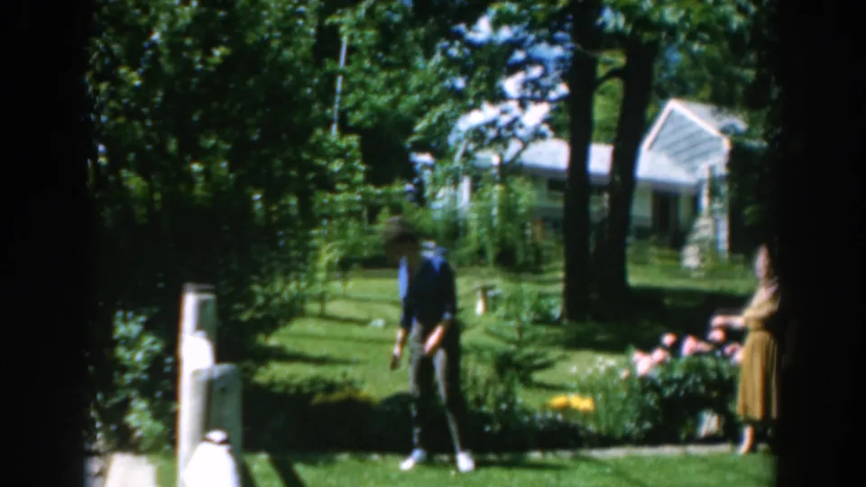 In 1962 Greece, a woman in a blue shirt stands on a lawn path while another woman is partly visible near flowers in a garden.