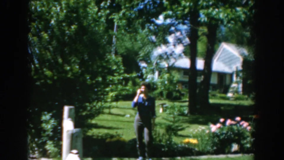 A woman in a blue shirt gestures or raises her hand in a leafy Greek garden, with flowers and a white house behind her.