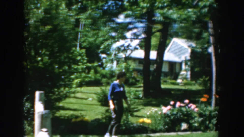 In the 1962 Greek home-movie scene, a woman in a blue shirt walks through a green garden beside flowering plants.