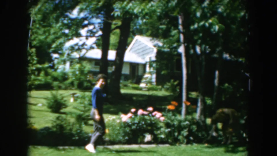 A woman in a blue shirt moves past a flower bed in a Greek garden, with trees and a white house visible in the background.