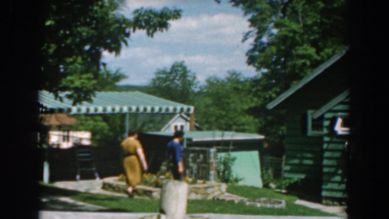 1962 Greece 8mm frame showing two people crossing a sunny garden near green-sided houses, a striped awning, trees, and flower beds.