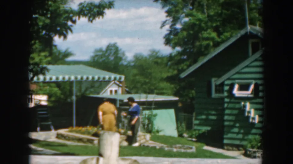 1962 Greece home-movie frame with a woman in yellow and a person in blue near a tended garden bed, green buildings, awnings, and trees.