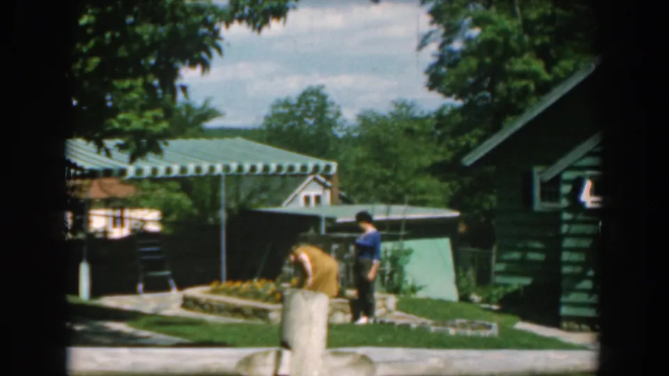 1962 Greece 8mm frame showing a woman bending at a garden bed as another person stands nearby, with green siding, lawn, trees, and awnings visible.
