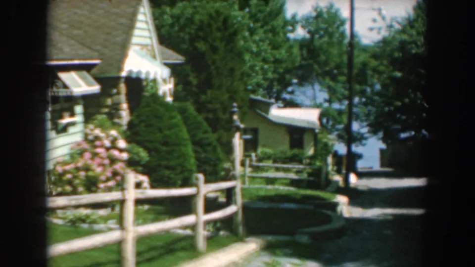 1962 Greece home-movie frame looking along a garden path with a wooden fence, green-painted structures, shrubs, trees, and possible water in the distance.