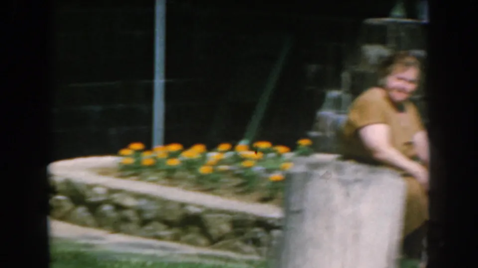 In 1962 Greece home-movie footage, a woman sits on a low garden wall near orange flowers and a shaded stone exterior.
