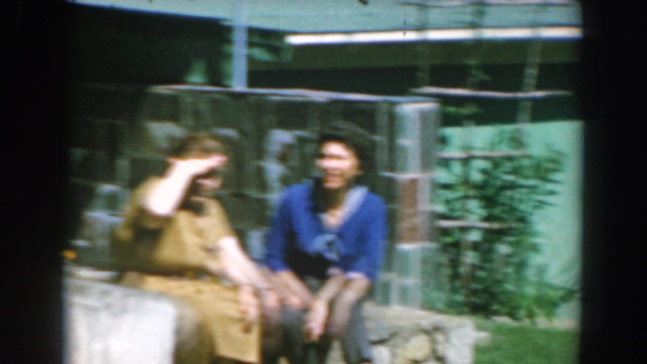 Two women sit on a low wall, one shielding her eyes in bright sun.