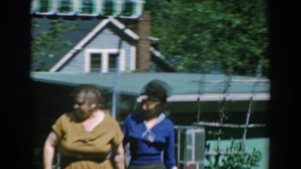 Two women move through the garden area, with a low wall, house roofline, chimney, and awning visible behind them.