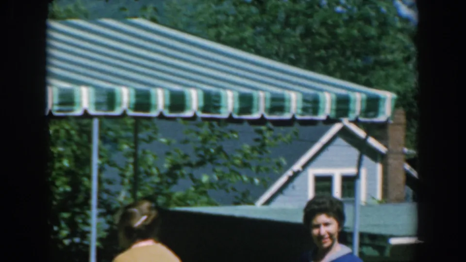 A striped awning, green roofline, and house exterior frame the garden scene as the women are partly visible below.