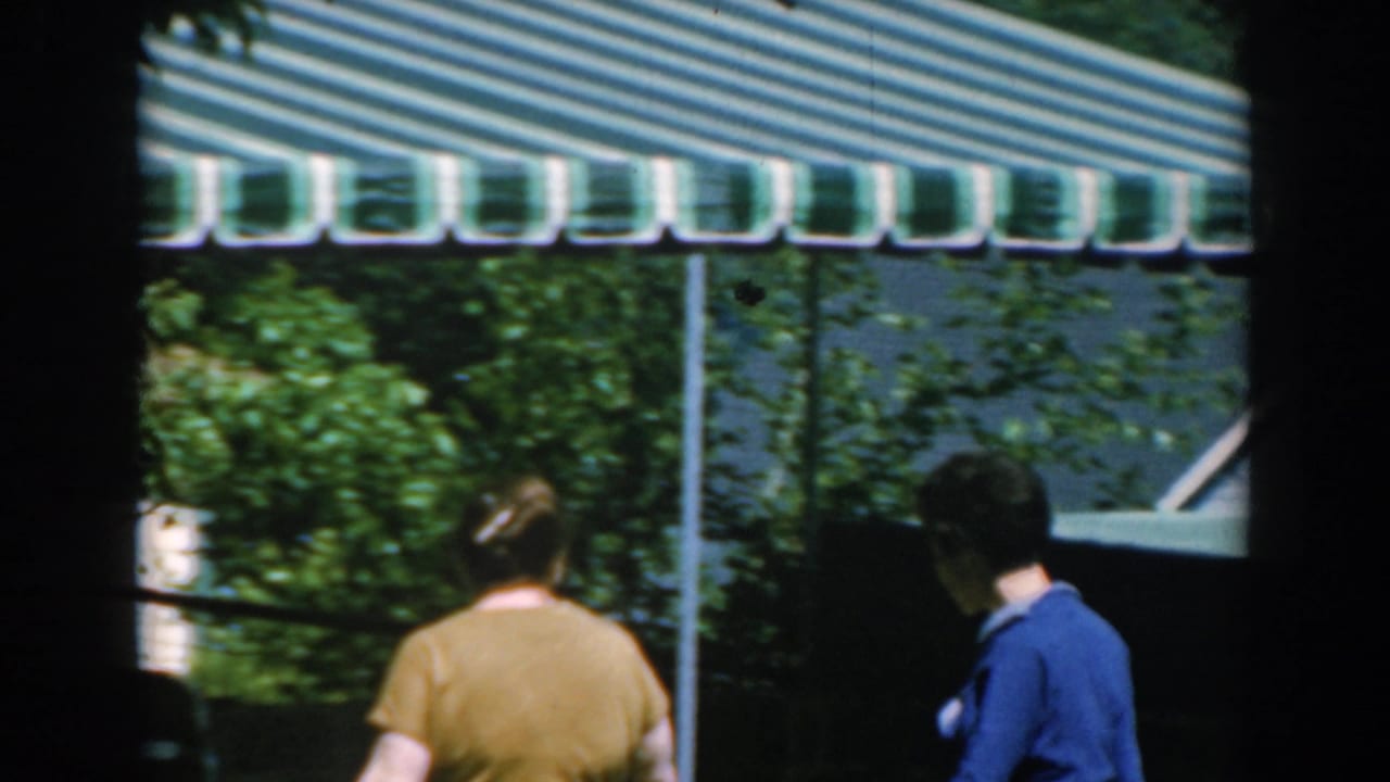 In 1962 Greece home-movie footage, two adult women walk away from the camera under a striped awning in a sunny garden setting.