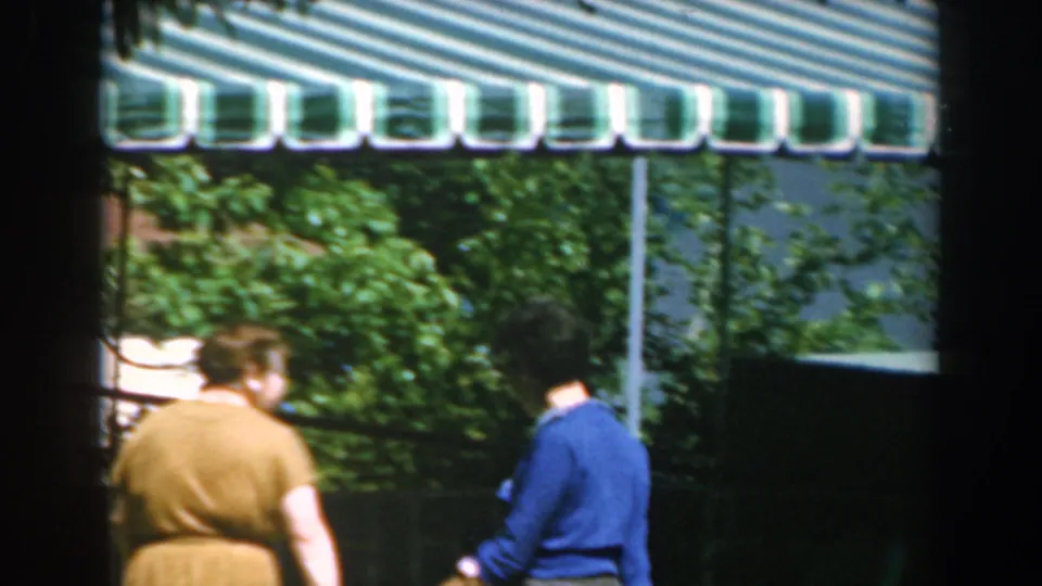 Two adult women in blue and yellow clothing move through a sunny Greek outdoor setting beneath a green and white awning.