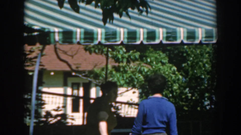 In the 1962 Greece clip, the women move through shadow below the awning while a house or terrace appears behind leafy branches.