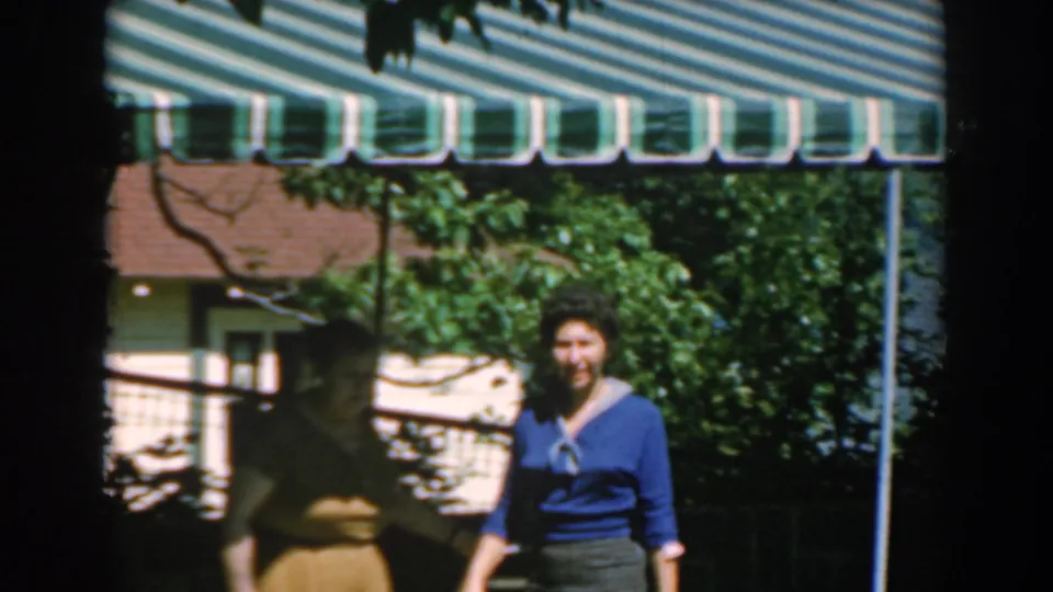 Near the end of the 1962 Greece home movie, the woman in blue turns toward the camera under the striped awning as another person passes in shade.