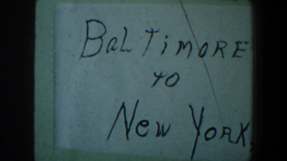 1960 8mm frame showing a handwritten Baltimore to New York travel note on pale paper with visible film borders.