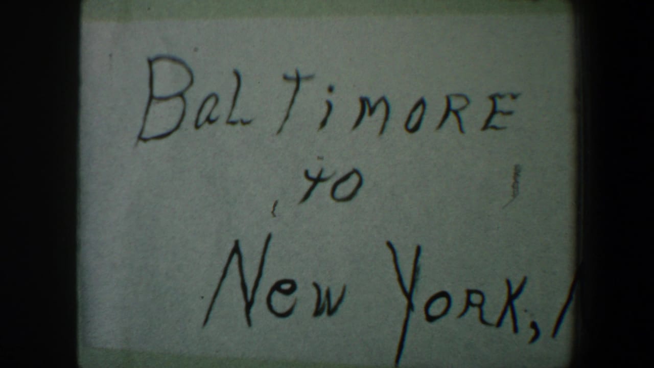 1960 8mm frame of a handwritten Baltimore to New York travel note, with soft focus, pale paper, and dark film border shadows.