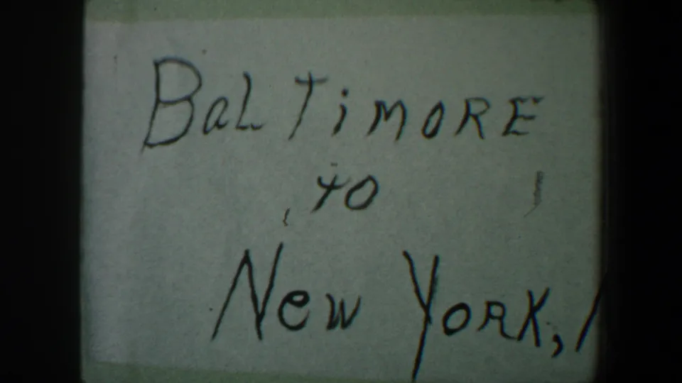 1960 8mm frame of a handwritten Baltimore to New York travel note, with soft focus, pale paper, and dark film border shadows.