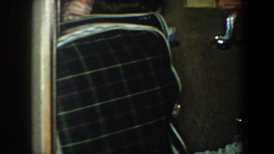 1960 New York City home-movie frame showing a large plaid suitcase in a dim interior hallway, with a person partly visible at the edge.