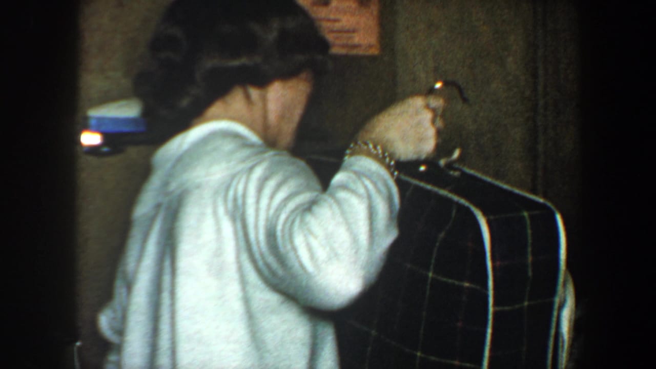 Woman in a white sweater holds a large plaid suitcase in a dim hallway.