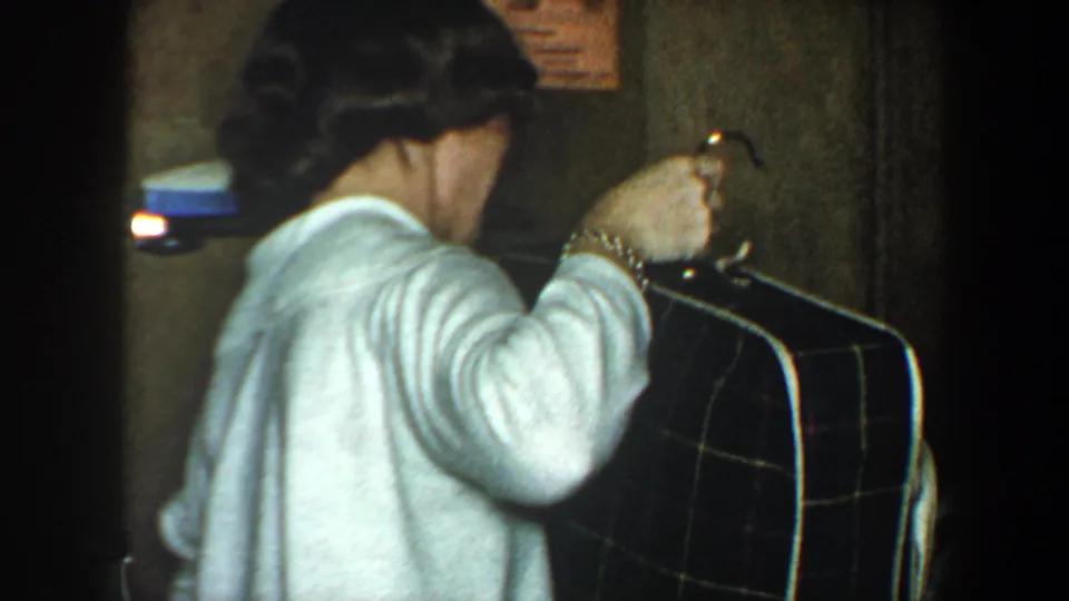 Woman in a white sweater and jewelry grips a large plaid suitcase in a dim New York City interior from 1960 home-movie footage.