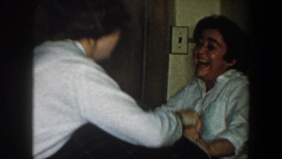 A laughing woman in a white blouse is seen with another person nearby in a tight 1960 New York City interior hallway scene.