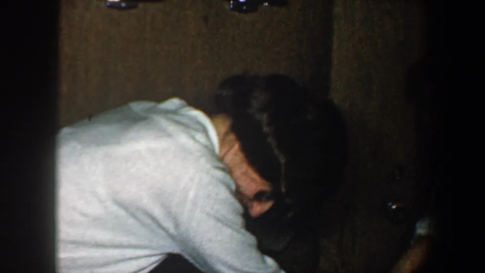 The woman bends over beside the plaid suitcase in a dim private interior, continuing the playful luggage struggle from the 1960 footage.