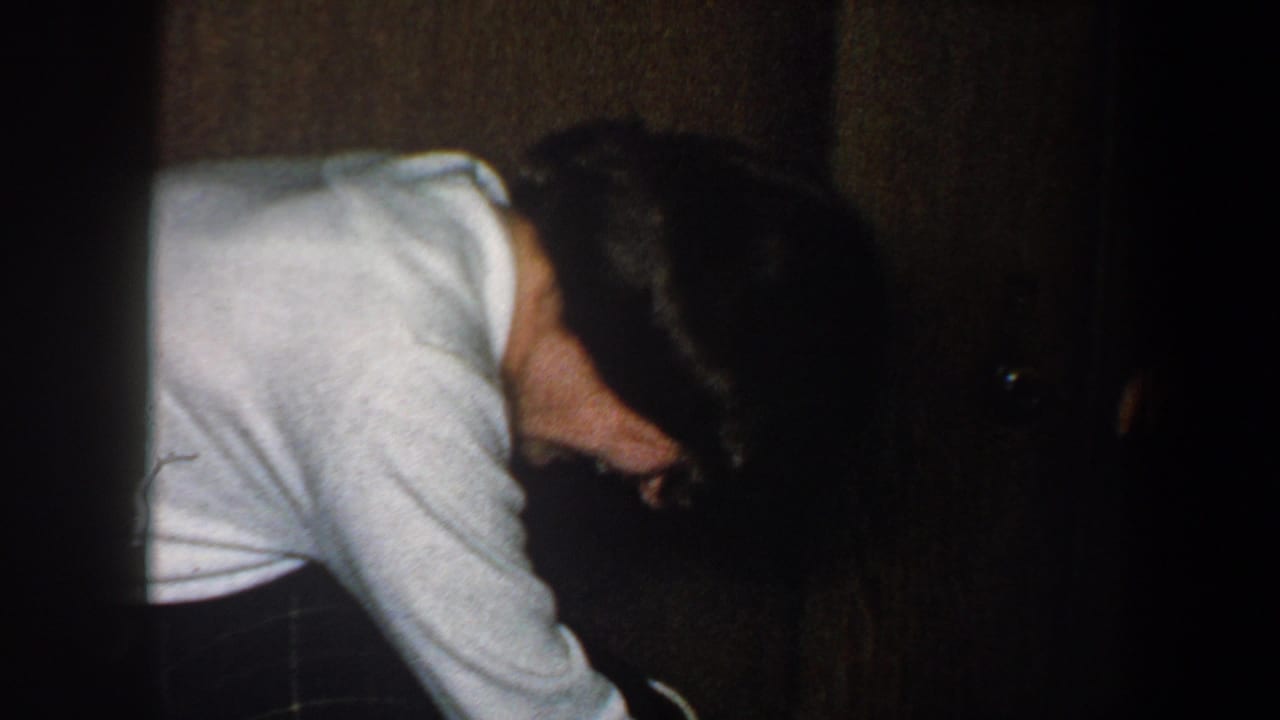 In 1960 New York City home-movie footage, an adult in a light shirt bends toward the floor inside a dark wood-paneled interior.