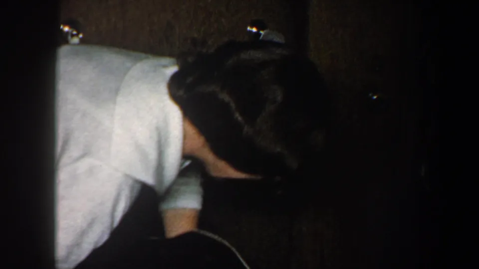 An adult in a light shirt leans forward in a dim interior, with another figure partly visible in shadow nearby.