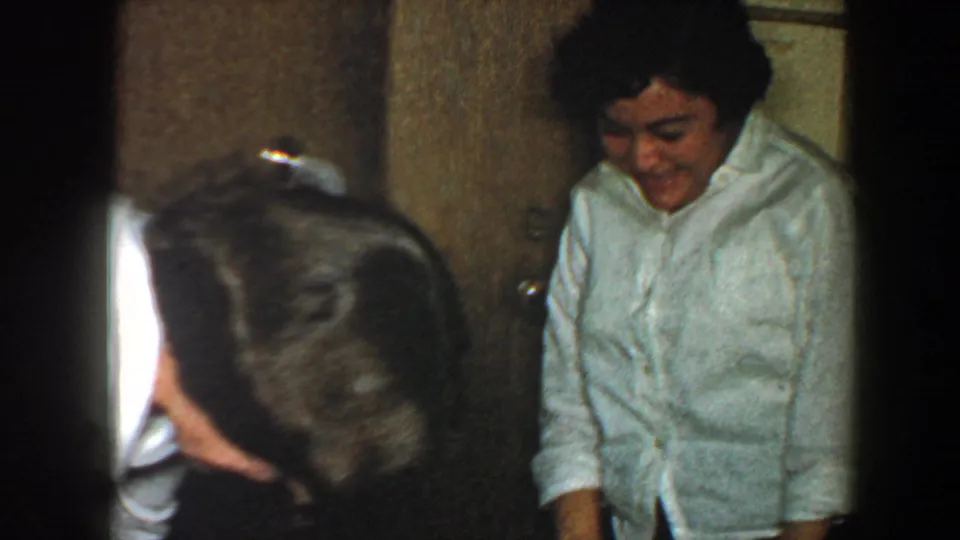 A woman in a light top stands against a wood-paneled wall while another adult bends in the foreground of the 1960 New York City home movie.
