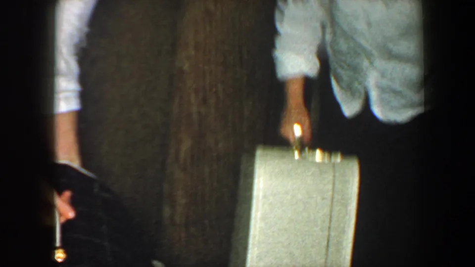 An adult reaches near a small light-colored suitcase in a dim wood-paneled interior, suggesting belongings being gathered from the floor.