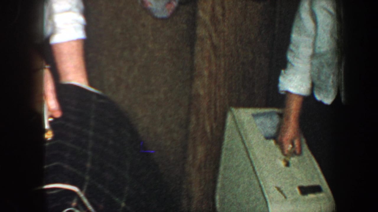 Partial adults in a dim hallway beside a hard-sided suitcase and wood door.