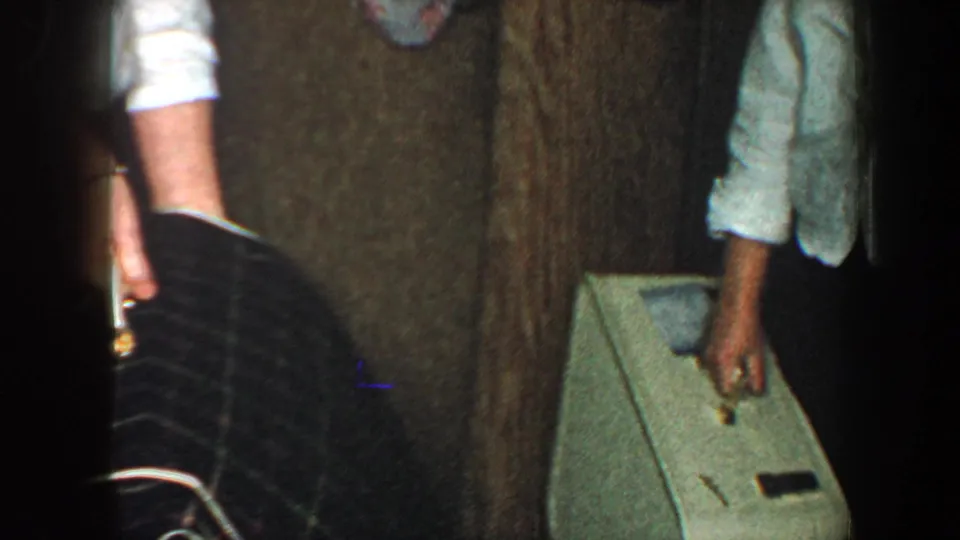 1960 New York City home-movie frame showing partial adults in a narrow hallway, with a light suitcase near a wood-paneled door.