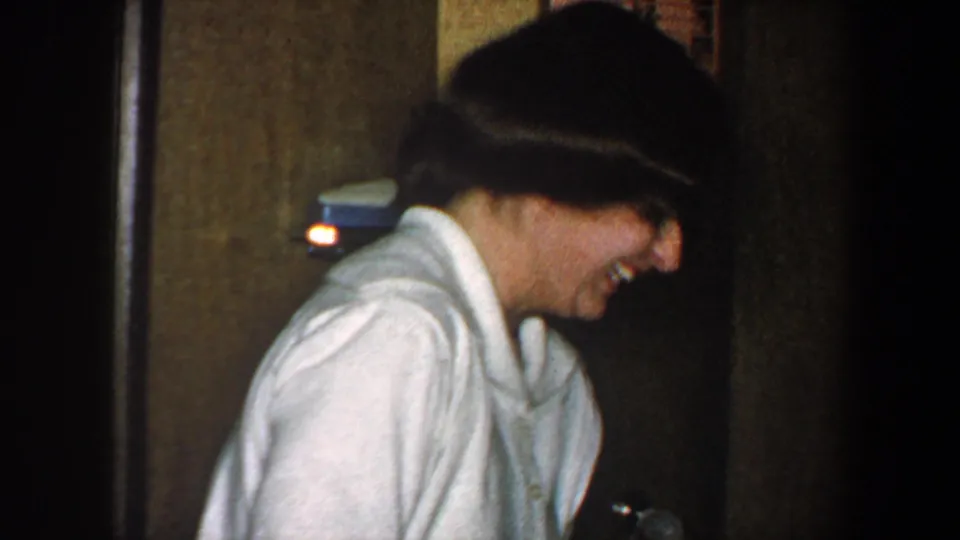 1960 New York City home-movie frame of a woman laughing in profile inside a dim private hallway with wood doors behind her.
