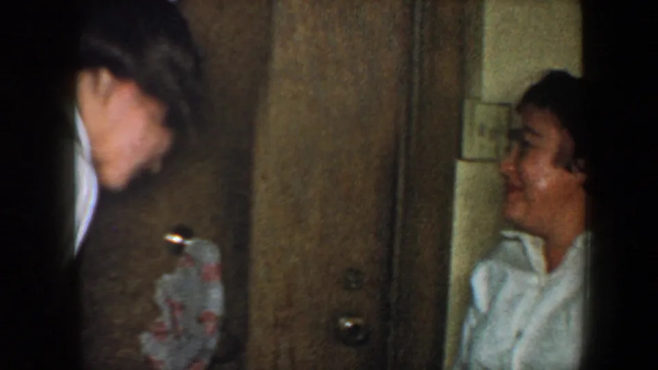 1960 New York City home-movie frame with two partially seen adults in a narrow hallway, one near a wall with a hanging patterned cloth.