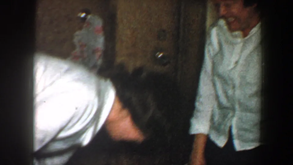 1960 New York City home-movie frame showing a woman bending forward with laughter in a hallway as another adult stands nearby.